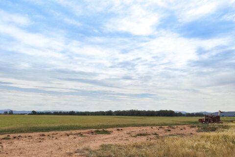 A vast field under a partly cloudy sky extends to the horizon, embodying a reimagine tradition. In the distance, a tractor moves near the edge of the field. The foreground features a blend of green grass and earthy terrain, while woodland and low hills line the horizon.