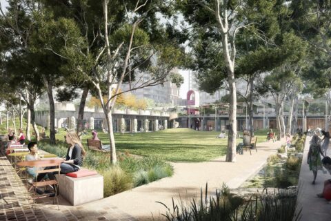 A lively scene unfolds at MacPark, Midtown's democratic heart. On the left, people are seated at a table, while others walk or relax on benches. To the right, children play on a playground. The setting is lush with trees and a grassy area; buildings are visible in the background.