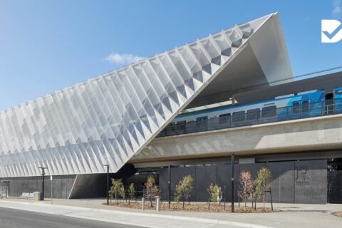 A modern train station, Reservoir Station, boasts a striking angular design with a white geometric facade. Elevated train tracks feature a blue train passing by. Below, minimalistic landscaping and young trees enhance the space. The image proudly displays the "Good Design Award Winner" logo.