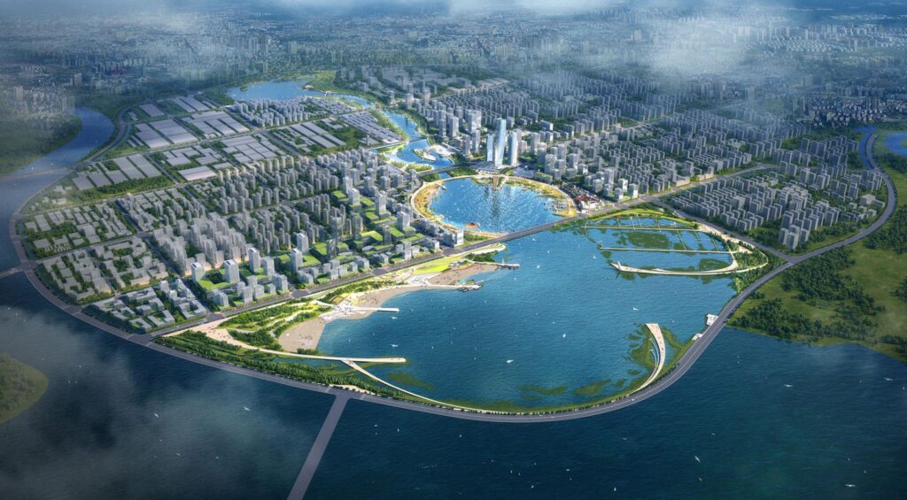 Aerial view of a modern, expansive urban development in Qingdao, surrounded by water and greenery. High-rise buildings cluster around central blue water bodies like Ruyi Lake, intersected by roads and bridges. Parks and green spaces are interspersed among residential and commercial structures, creating a harmonious environment.