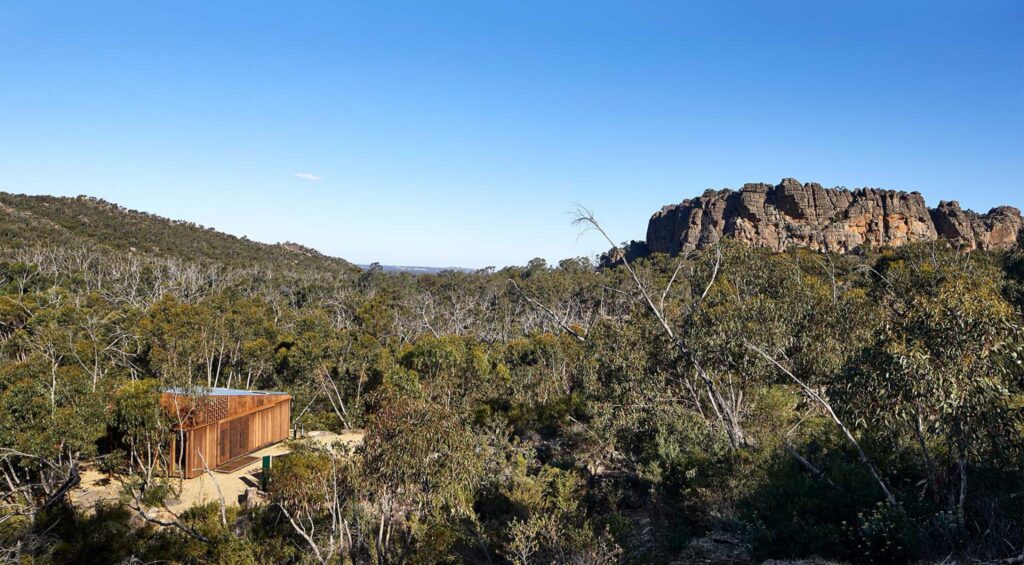 A small wooden cabin is nestled in a dense forest with a clear blue sky overhead. In the background, rugged, rocky cliffs rise above the tree line, creating a dramatic and picturesque landscape. This Australian eco-tourism retreat takes visitors to new heights of natural beauty and tranquility.