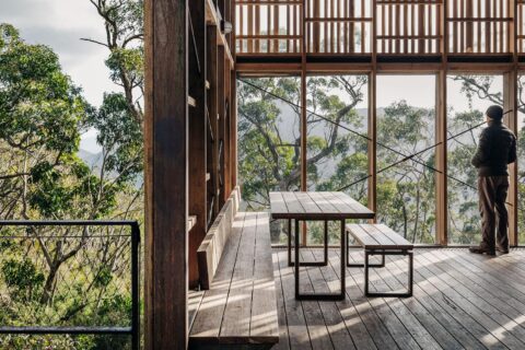 A person stands by the large windows of a rustic, wooden modern cabin overlooking the dense forest of the Grampians Peaks Trail. The interior features minimalist furniture, including a rectangular table and benches. Natural light streams in through the tall windows, creating a serene atmosphere.