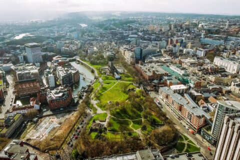 Aerial view of a cityscape featuring a large green park in the center surrounded by various buildings, streets, and waterways. The park includes winding paths and trees, with new homes dotted around. The city expands outward with a mix of modern and traditional architecture.