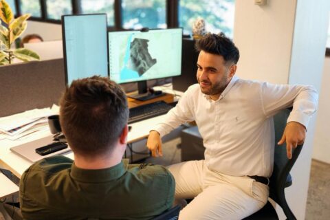 Two men are having a discussion at a workstation with dual monitors. One man, in a white shirt, is sitting in a chair facing the other man in a green shirt. The monitors display what seems to be topographical data. The Biourbanism Lab, with its focus on prosperity by design, sets the office backdrop.
