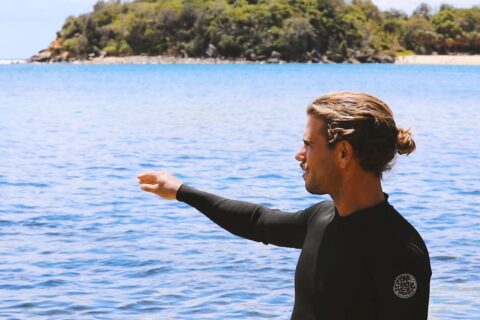 A person with long hair tied in a bun, wearing a black wetsuit, stands near the ocean, pointing towards the water. In the background, there is a scenic view of a lush green island and the clear blue sea under a partly cloudy sky—showcasing their commitment to ocean conservation.