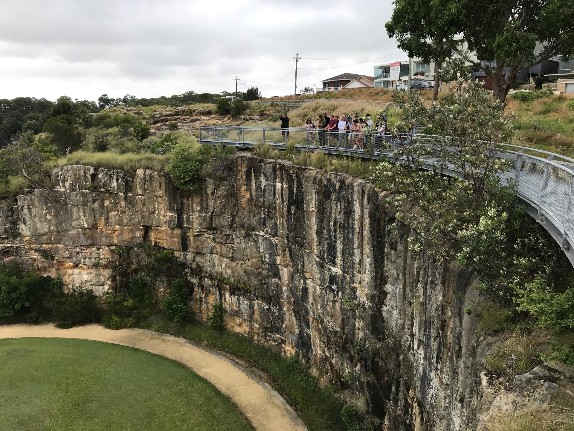 A group of people stands on a curved metal viewing platform overlooking a deep limestone quarry with green grass at the bottom. The surrounding landscape, once a former BP site, is grassy with scattered trees and houses visible in the background under a cloudy sky.