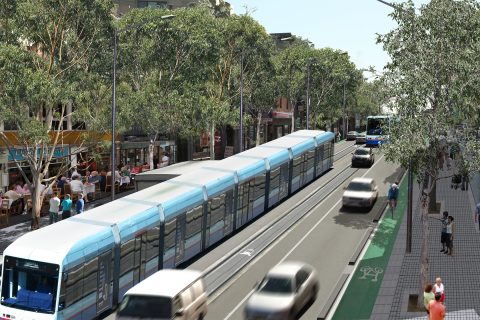 A modern light rail train travels down a tree-lined city street, flanked by moving cars on Parramatta Road and a dedicated bike lane. Pedestrians walk on wide sidewalks, with outdoor dining on the left. The scene is vibrant and sunny, reflecting urban public transportation and community life.