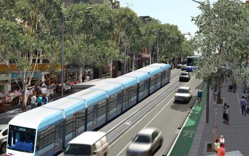 A modern light rail train travels down a tree-lined city street, flanked by moving cars on Parramatta Road and a dedicated bike lane. Pedestrians walk on wide sidewalks, with outdoor dining on the left. The scene is vibrant and sunny, reflecting urban public transportation and community life.