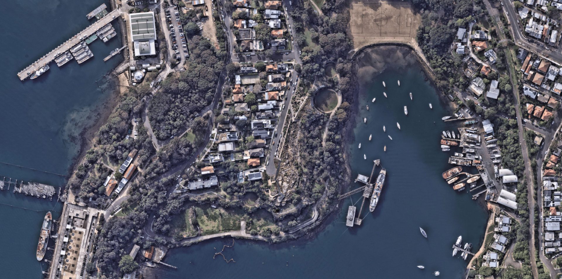 Aerial view of a coastal residential area with numerous houses surrounded by trees. Former BP Site Park lies within, adding greenery to the landscape. There are several boats anchored in the water along the coastline. Roads weave through the area, and there are docks with boats, including some larger vessels.