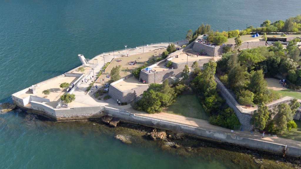 Aerial view of a coastal fortress at Ballast Point Park, with stone walls extending into the water. The structure features multiple levels with trees and pathways. Visitors are dispersed throughout the grounds. The surrounding water is calm, and the sky is clear.