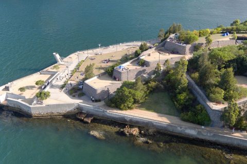 Aerial view of a coastal fortress at Ballast Point Park, with stone walls extending into the water. The structure features multiple levels with trees and pathways. Visitors are dispersed throughout the grounds. The surrounding water is calm, and the sky is clear.