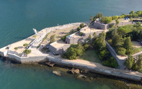 Aerial view of a coastal fortress at Ballast Point Park, with stone walls extending into the water. The structure features multiple levels with trees and pathways. Visitors are dispersed throughout the grounds. The surrounding water is calm, and the sky is clear.