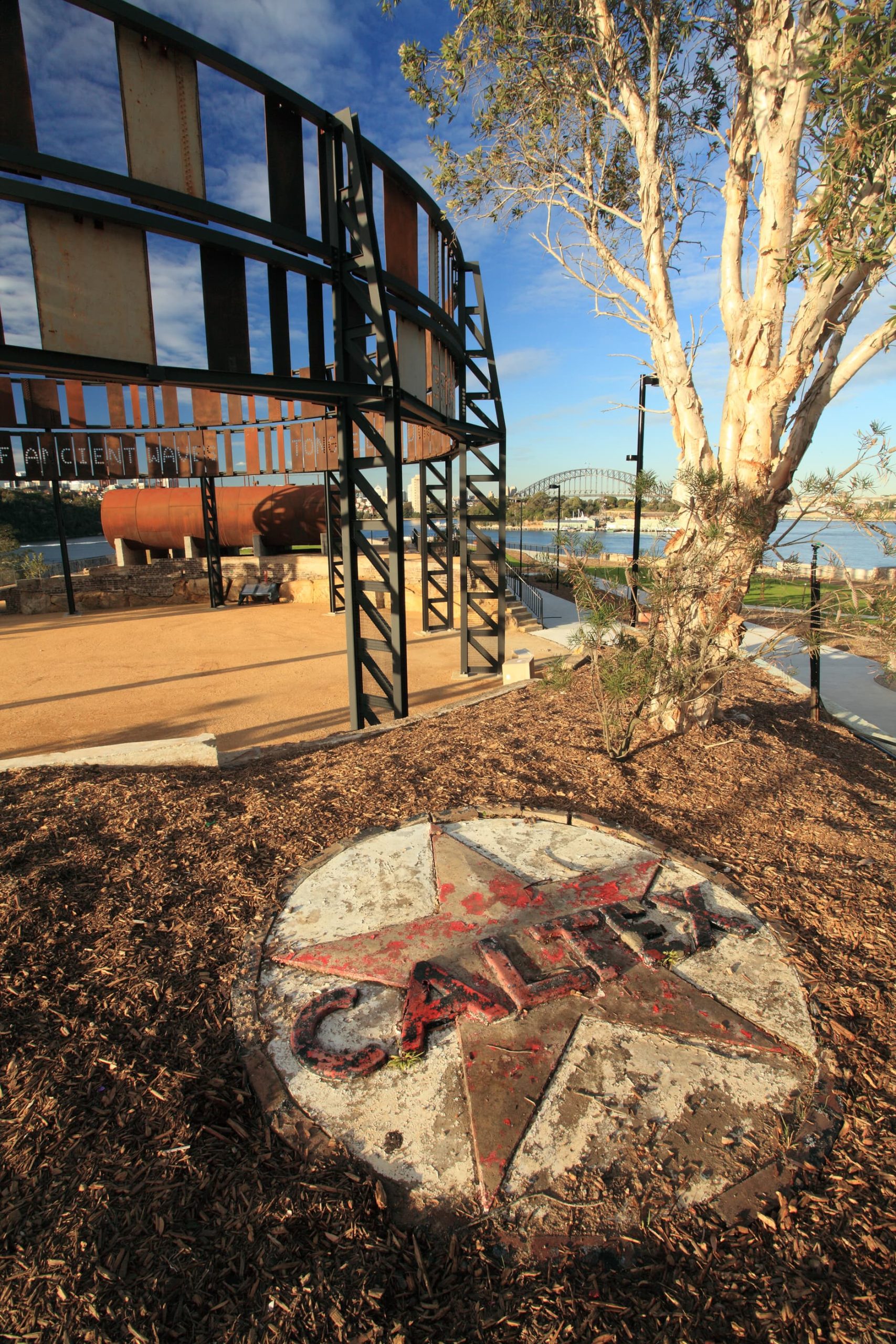 An outdoor area at Ballast Point Park features a large weathered Texaco CA-LIFE star logo embedded in the ground at the forefront. A tree stands nearby, and in the background, industrial structures and a bridge stretch under a clear blue sky.