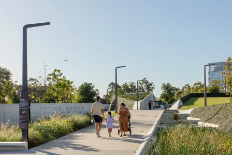 A family of four walks along a paved path in a modern urban park with greenery, trees, and a pond. Street lamps and a sign reading "DRYING GREEN" are visible. One family member pushes a stroller, while others hold hands under a clear blue sky. An airplane is in the distant sky.