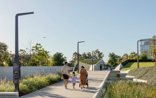 A family of four walks along a paved path in a modern urban park with greenery, trees, and a pond. Street lamps and a sign reading "DRYING GREEN" are visible. One family member pushes a stroller, while others hold hands under a clear blue sky. An airplane is in the distant sky.