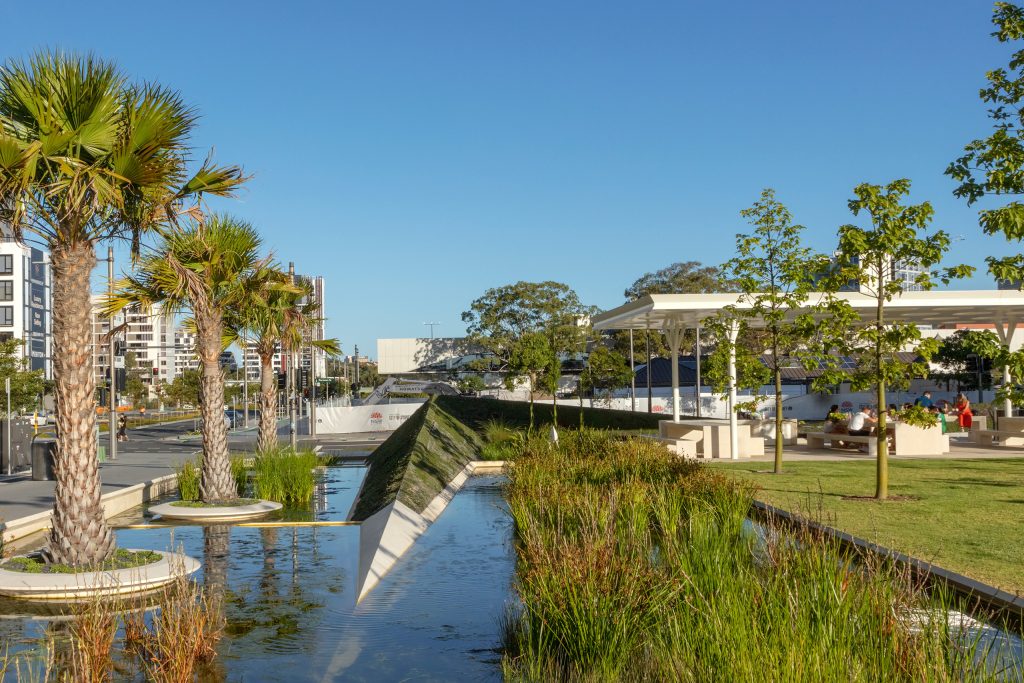 The Drying Green is a modern park with palm trees and a small stream running through it. The park includes landscaped grass areas, a pavilion with people sitting at tables, and modern buildings in the background. The sky is clear and blue.