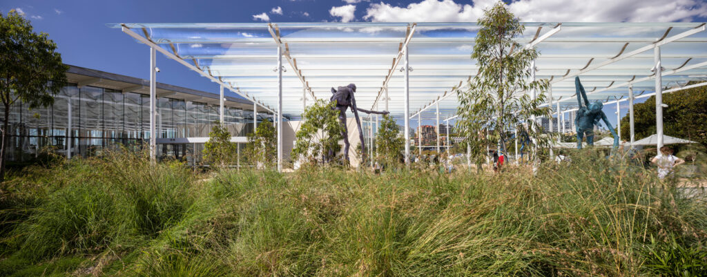 Modern outdoor pavilion with a transparent roof structure housing contemporary sculptures, surrounded by tall grasses and trees. A glass-walled building is visible on the left. A sculpture of a human figure stands in the foreground, while another in blue tones is positioned further behind. Sydney's skyline forms a distant backdrop.