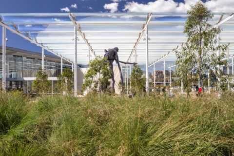 Modern outdoor pavilion with a transparent roof structure housing contemporary sculptures, surrounded by tall grasses and trees. A glass-walled building is visible on the left. A sculpture of a human figure stands in the foreground, while another in blue tones is positioned further behind. Sydney's skyline forms a distant backdrop.