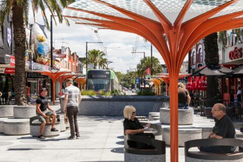 A bustling urban street scene on Acland Street features people sitting under modern, orange tree-shaped structures. Shops and palm trees line both sides of the street, with a green tram in the distance. The sky is partly cloudy, capturing the lively essence of St Kilda.