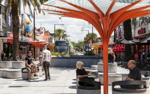 A bustling urban street scene on Acland Street features people sitting under modern, orange tree-shaped structures. Shops and palm trees line both sides of the street, with a green tram in the distance. The sky is partly cloudy, capturing the lively essence of St Kilda.