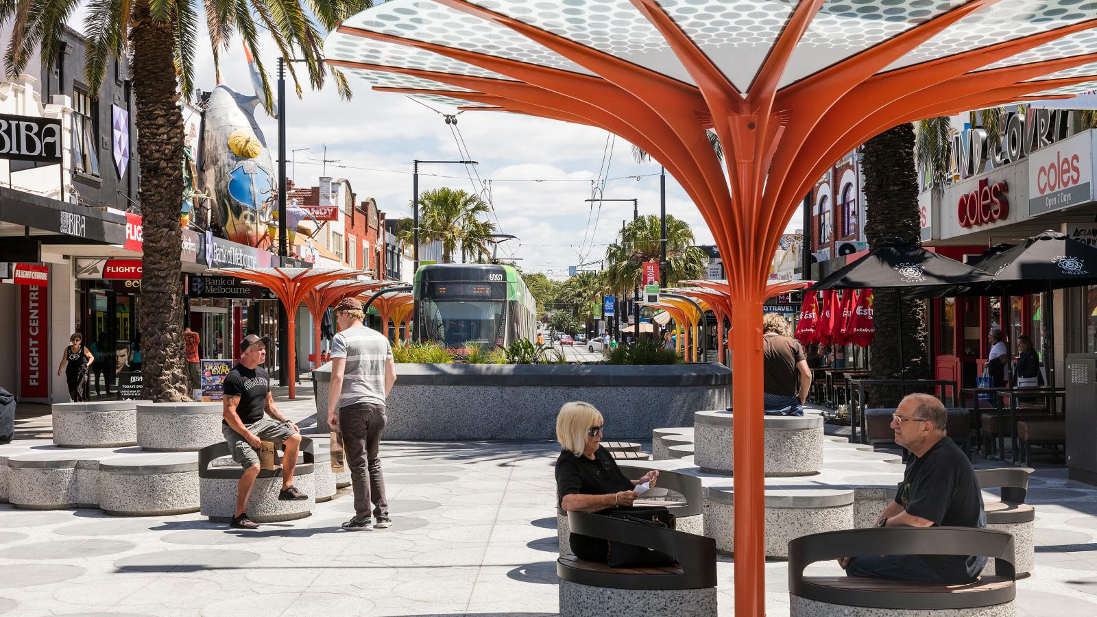 A bustling urban street scene on Acland Street features people sitting under modern, orange tree-shaped structures. Shops and palm trees line both sides of the street, with a green tram in the distance. The sky is partly cloudy, capturing the lively essence of St Kilda.