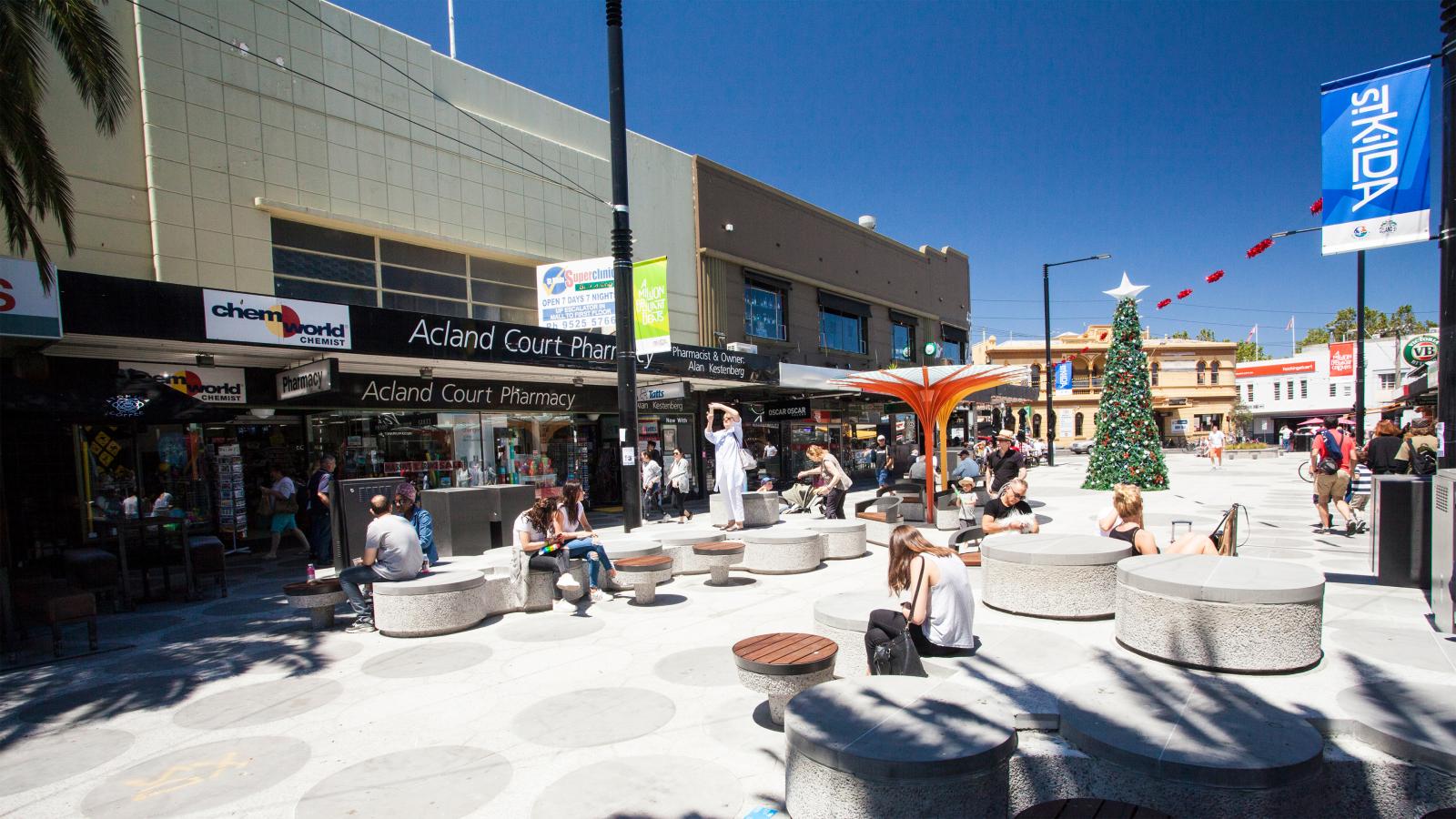 People are sitting on circular concrete benches in a lively plaza in St Kilda, with shops like Acland Court Pharmacy and Chemist World. A large Christmas tree and a flower-shaped structure are also visible. Red and blue shop signs and flags adorn Acland Street against a clear blue sky.