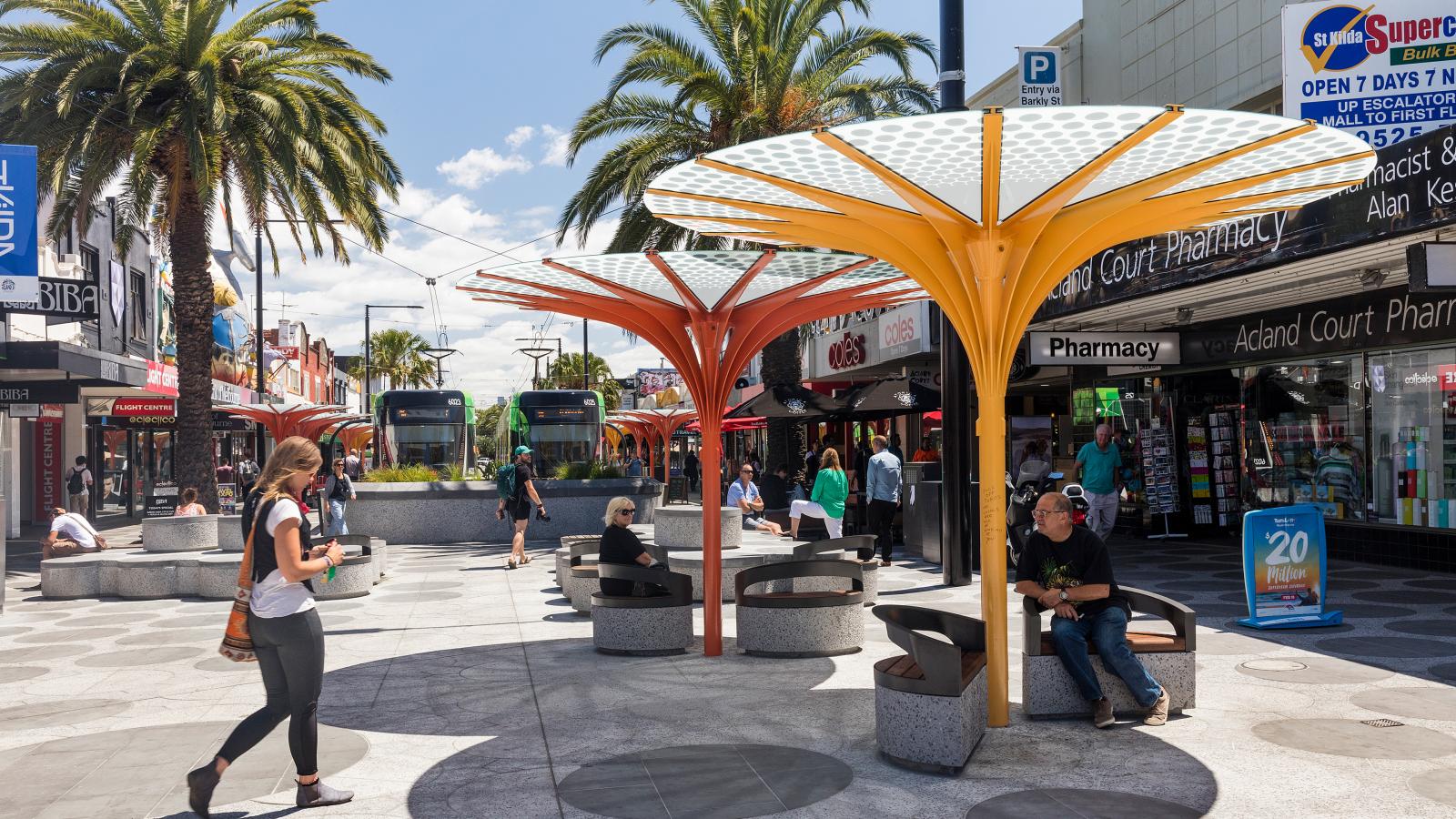 A bustling urban street scene on Acland Street, St Kilda, featuring people walking and sitting beneath modern, umbrella-shaped shade structures. In the background, two trams are visible, and shops including a pharmacy and a convenience store line the street. Palm trees add a tropical vibe.