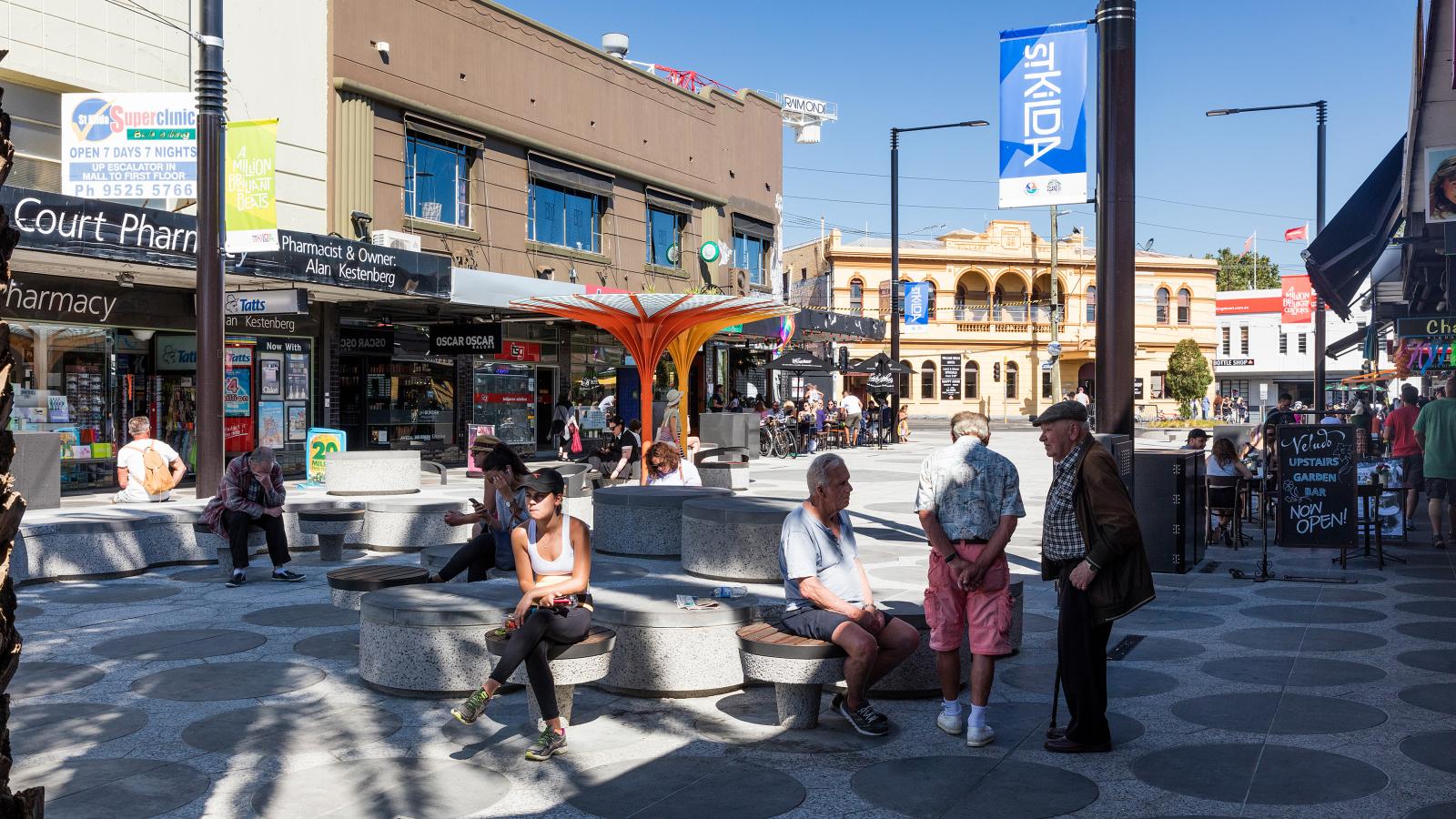 A busy outdoor commercial plaza on Acland Street, St Kilda, features people sitting on circular concrete benches and walking around. Various shops, including a pharmacy and convenience store, line both sides of the street. An orange modern art structure stands prominently in the middle of the plaza.