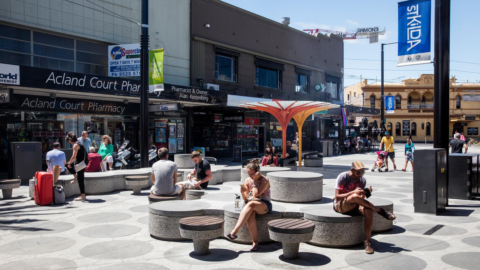 A busy urban plaza on Acland Street features people sitting and socializing on modern circular benches. The backdrop includes various shops, a pharmacy, and a distinctive mushroom-shaped canopy over one bench. Pedestrians move through the area, some with luggage or shopping bags.
