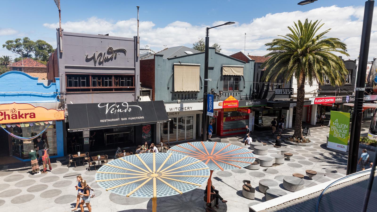 A lively urban street scene on Acland Street in St Kilda features shops, cafes, and restaurants with colorful geometric-patterned umbrellas. Pedestrians stroll along the pavement while others sit at outdoor tables. Palm trees and bright signage enhance the vibrant atmosphere on a sunny day.