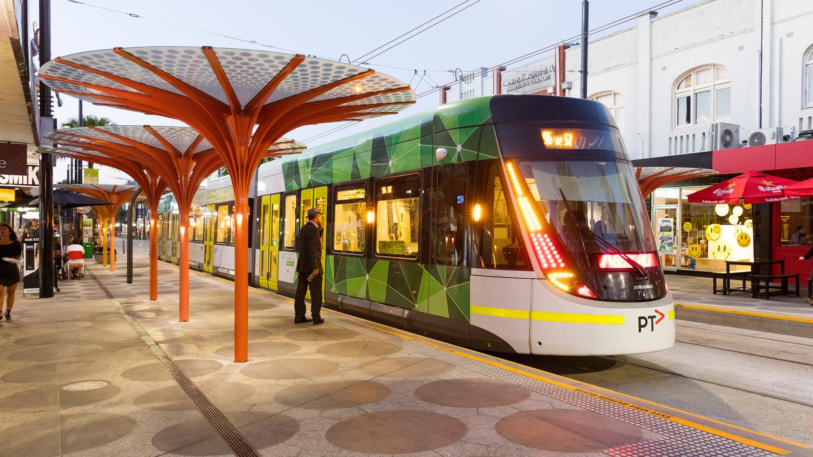 A modern tram with a green, geometric design is stopped at a platform in an urban area on St Kilda’s Acland Street. The station features unique, orange, umbrella-like structures. A person in a black jacket is stepping onto the tram. Nearby are storefronts and a red awning with additional seating.