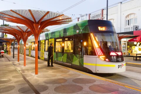 A modern green and white tram is stopped at a station with distinctive orange, tree-shaped shelters. A person is boarding the tram, illuminated and displaying "59A" on its front. Nearby sectors feature shops and cafes with bright signs.