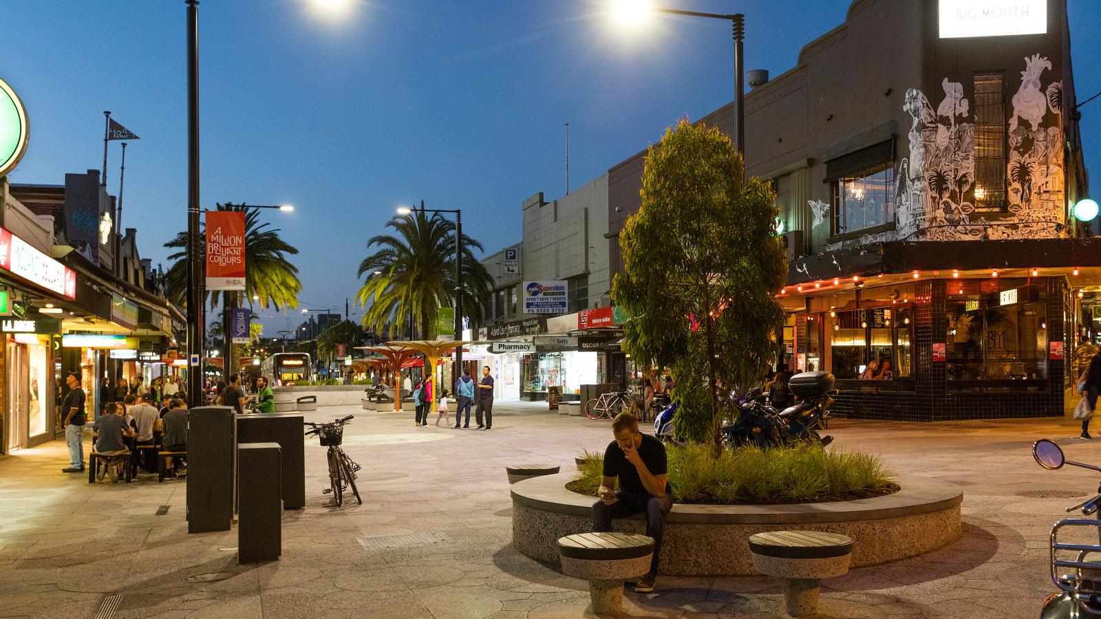 A bustling city street at dusk with shops, cafes, and restaurants. People sit and walk along the pedestrian area of Acland Street, illuminated by streetlights. A person sits on a bench near a tree in the foreground. Palm trees and modern buildings line the background in St Kilda.