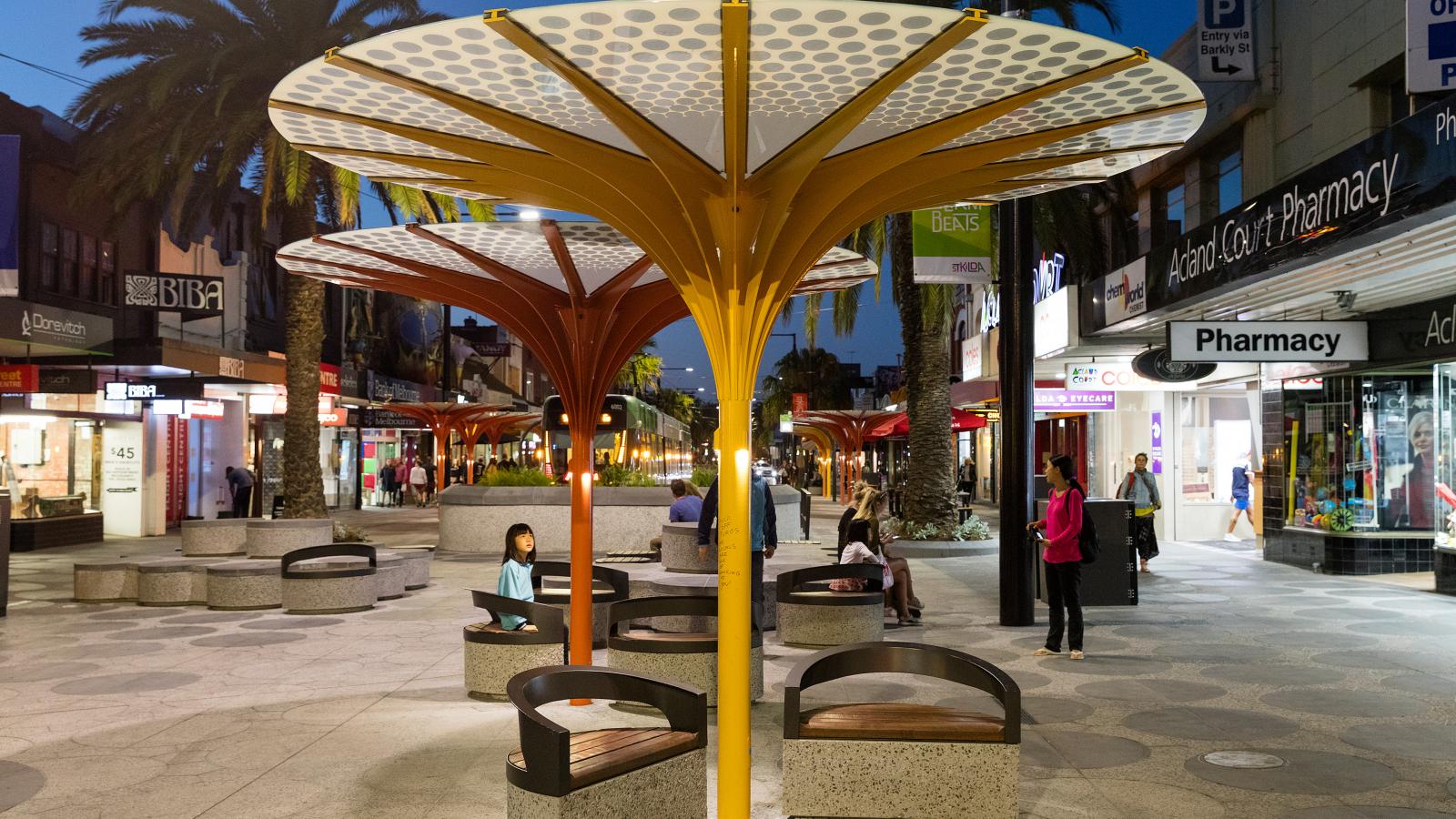 A vibrant, well-lit city street in the evening with modern umbrella-shaped structures providing shade over circular benches. People are seen walking and sitting on the benches. Shops and stores, including a pharmacy, line both sides of Acland Street in St Kilda.