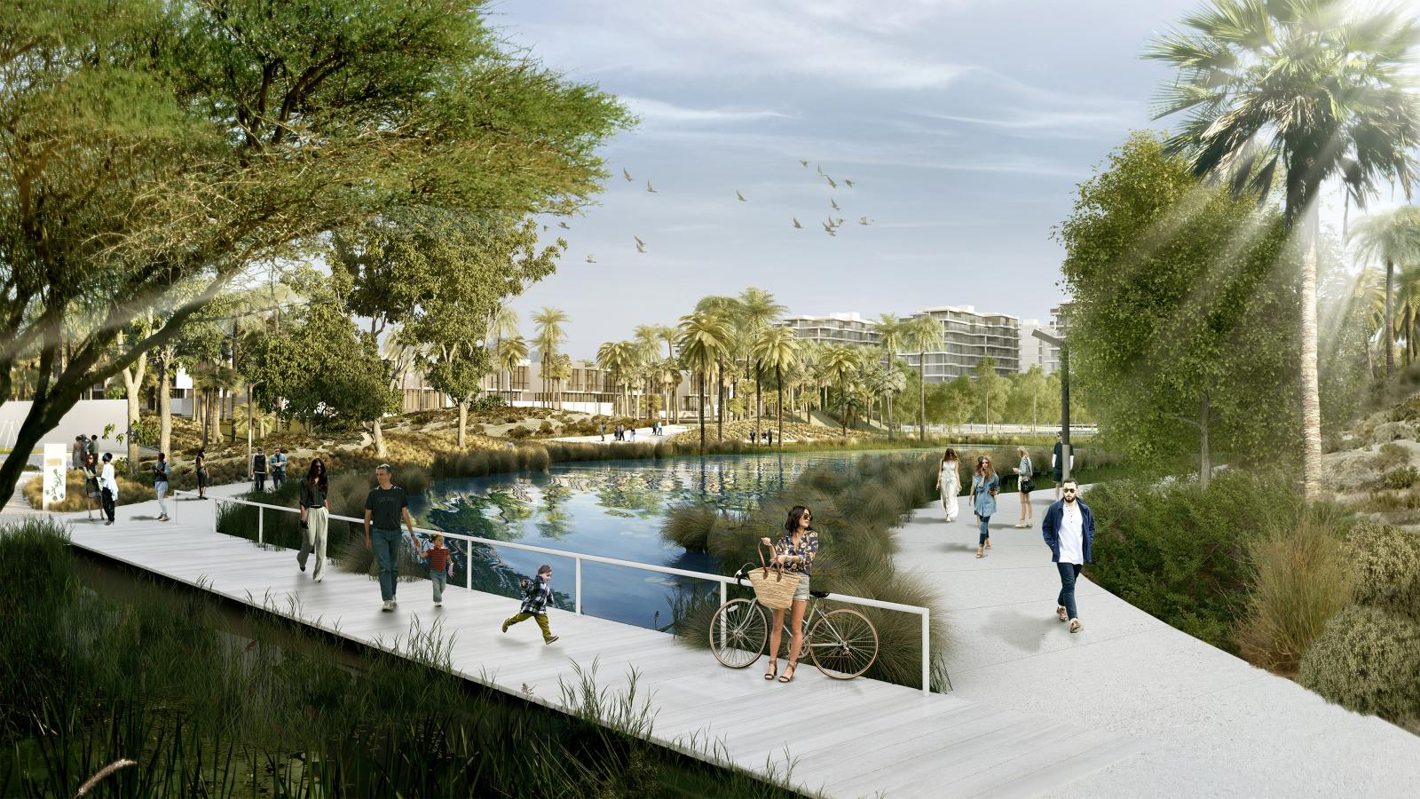 A serene Akoya Park scene with people walking and cycling along a boardwalk beside a calm water body. Tall trees and lush greenery surround the area. Birds fly overhead, and modern buildings are visible in the background under a clear, sunny sky.