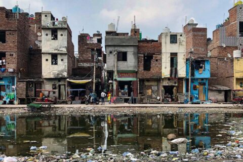 A row of aged, multi-story buildings stands closely packed along a street. The structures are in various states of disrepair. In front, a large puddle filled with garbage and debris reflects the buildings. People and a rickshaw are visible in the scene, highlighting the importance of water security initiatives like AIWASI.