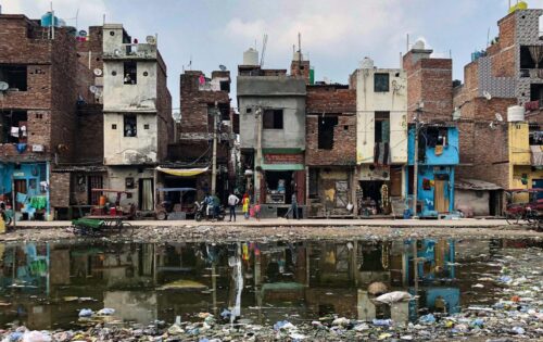 A row of aged, multi-story buildings stands closely packed along a street. The structures are in various states of disrepair. In front, a large puddle filled with garbage and debris reflects the buildings. People and a rickshaw are visible in the scene, highlighting the importance of water security initiatives like AIWASI.