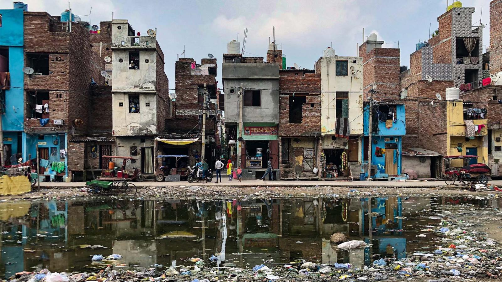 A row of aged, multi-story buildings stands closely packed along a street. The structures are in various states of disrepair. In front, a large puddle filled with garbage and debris reflects the buildings. People and a rickshaw are visible in the scene, highlighting the importance of water security initiatives like AIWASI.