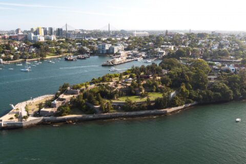 Aerial view of a lush, green peninsula extending into a body of water, surrounded by sailboats and yachts. The shoreline is dotted with parks like Ballast Point Park and residential areas, while a cityscape with tall buildings and bridges is visible in the background under a clear sky.