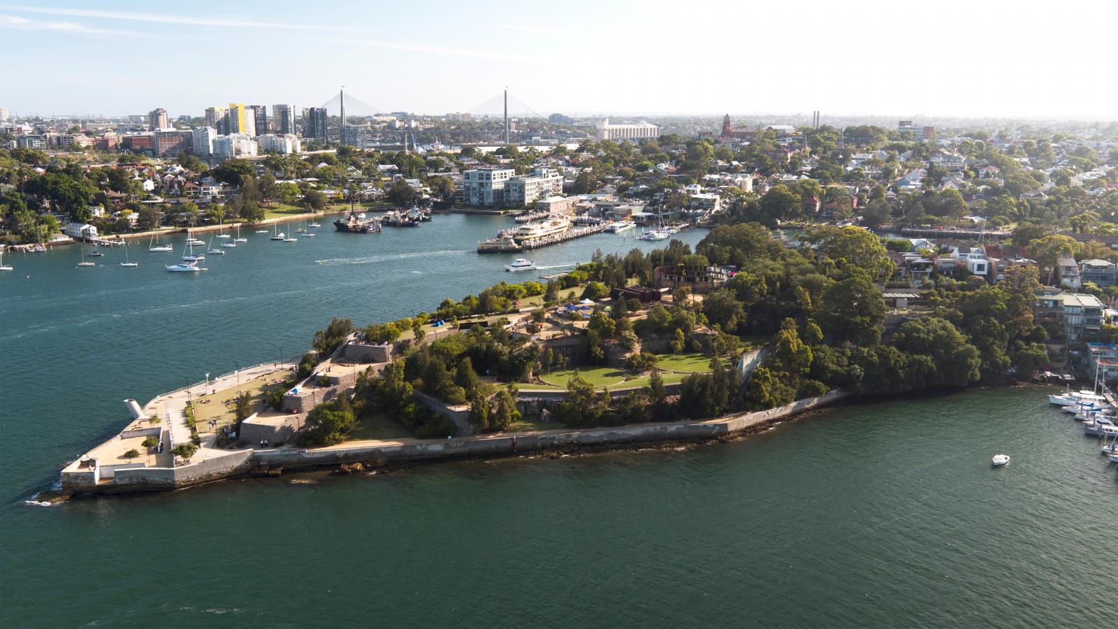 Aerial view of a lush, green peninsula extending into a body of water, surrounded by sailboats and yachts. The shoreline is dotted with parks like Ballast Point Park and residential areas, while a cityscape with tall buildings and bridges is visible in the background under a clear sky.