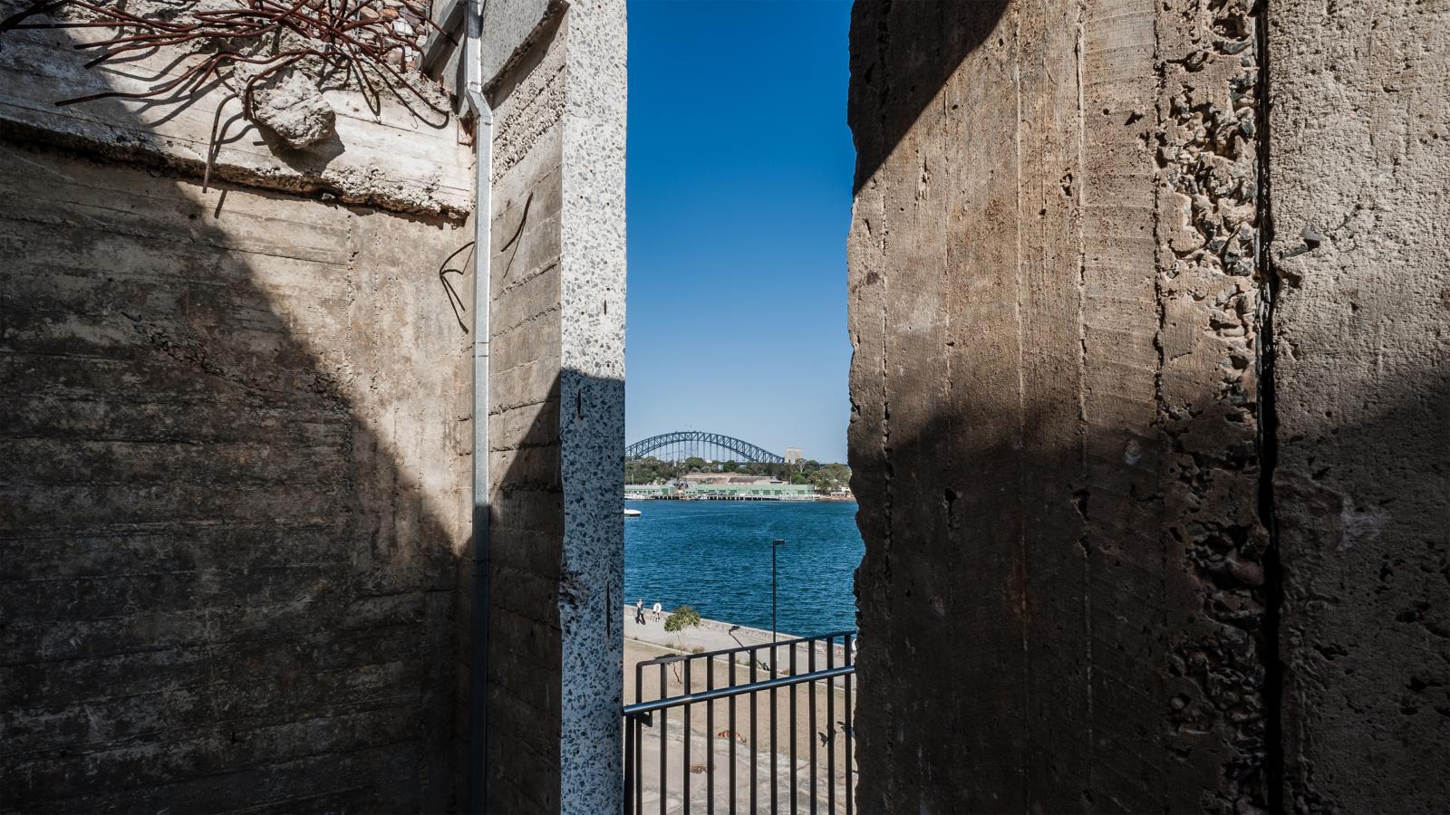 A view of a bridge framed by tall, textured concrete walls in Ballast Point Park. Between the walls, there's a body of water with a boat and part of a railing in the foreground. The sky is clear and blue.