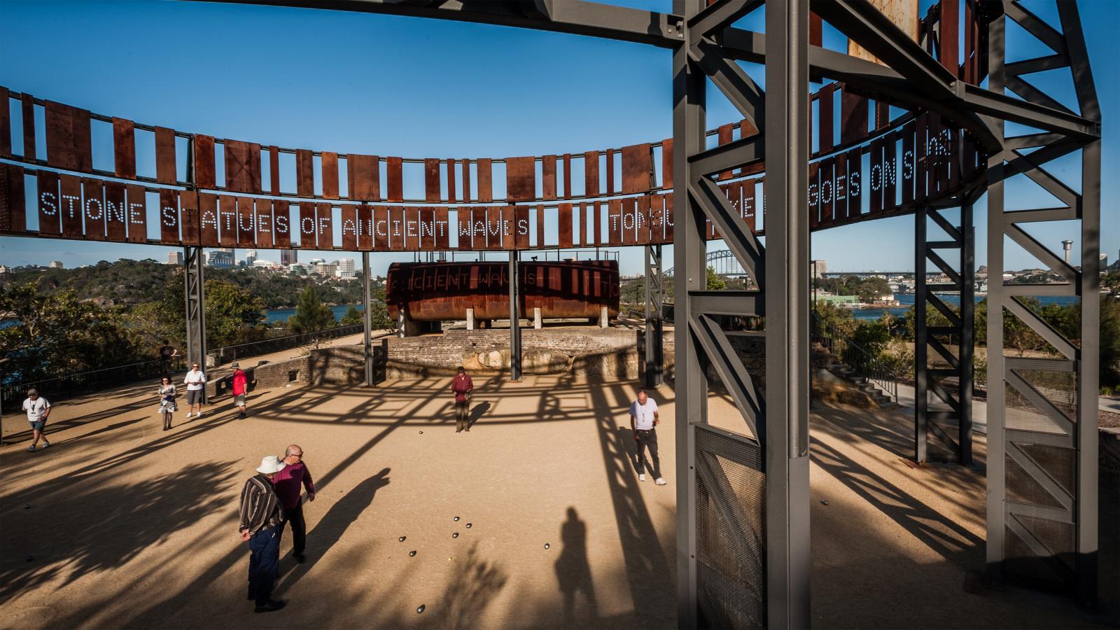 Visitors walk around the Westpac War Memorial on top of Mt. Coot-tha in Brisbane, reminiscent of Ballast Point Park. The memorial features towering steel structures, a circular arrangement partially inscribed with text, and offers scenic views of the surrounding landscape. Shadows of the structure fall on the ground.