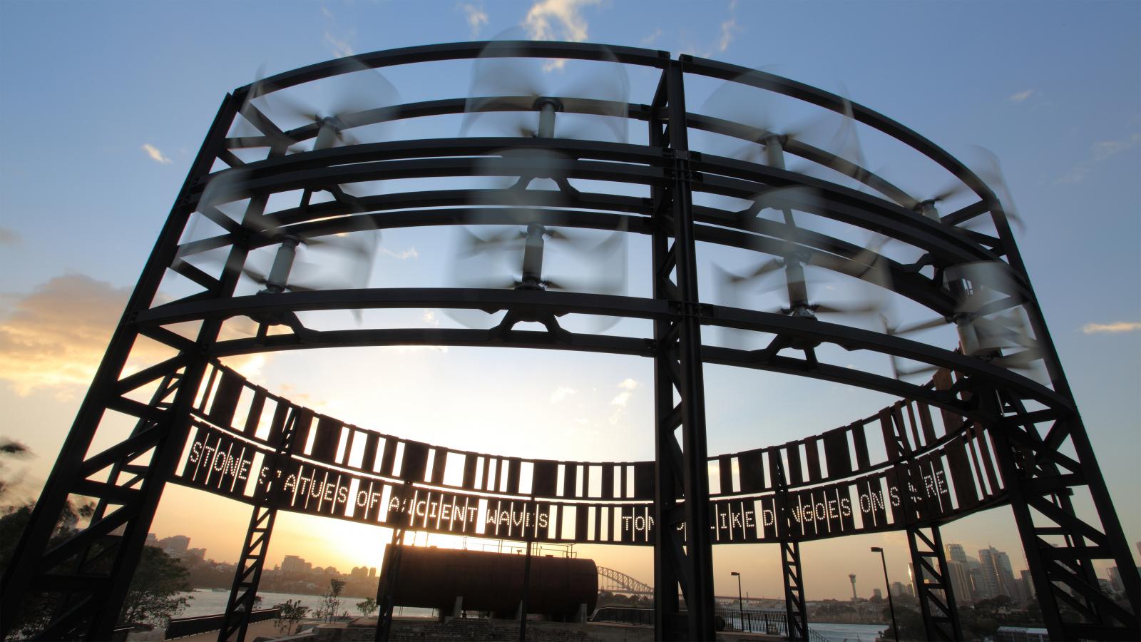 A circular kinetic light sculpture with spinning blades stands against a twilight sky in Ballast Point Park. The structure has multiple rows and appears to be in motion, creating a dynamic and captivating visual. The sun is setting in the background, casting a warm glow over the park.