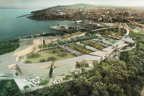 Aerial view of the sprawling Bandirma Park complex with modern architectural structures surrounded by greenery and gardens, adjacent to a large body of water. The cityscape extends into the background, with hills and buildings visible in the distance, creating an inviting competition ground.