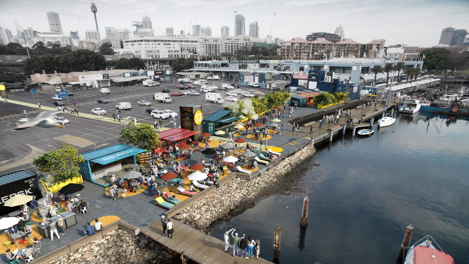 A bustling waterfront scene features people dining and socializing at outdoor tables along a promenade lined with colorful umbrella-shaded seating areas and food stalls. Adjacent to the lively promenade is a parking lot with parked cars and city buildings in the background.