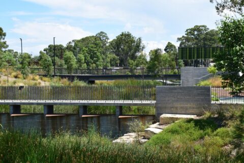 A scenic view of Blacktown Showground park with a bridge crossing over a waterway. The area is surrounded by lush greenery and tall trees. The sky is partially cloudy, and the park appears peaceful and well-maintained with a mix of natural and man-made elements.