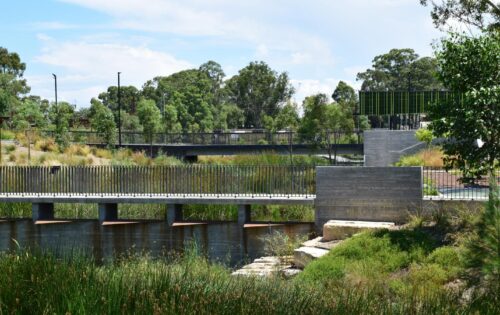 A scenic view of Blacktown Showground park with a bridge crossing over a waterway. The area is surrounded by lush greenery and tall trees. The sky is partially cloudy, and the park appears peaceful and well-maintained with a mix of natural and man-made elements.