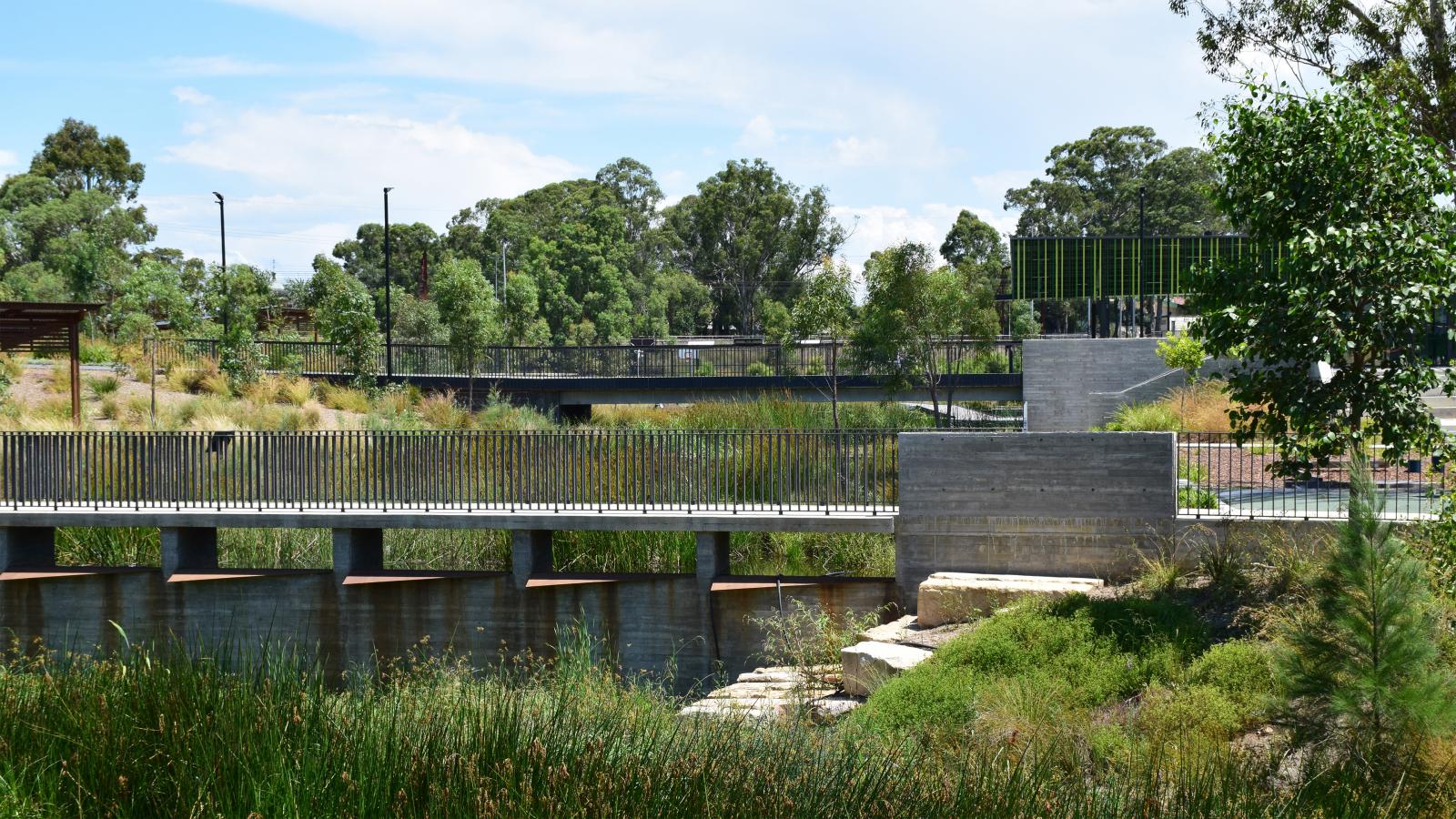 A scenic view of Blacktown Showground park with a bridge crossing over a waterway. The area is surrounded by lush greenery and tall trees. The sky is partially cloudy, and the park appears peaceful and well-maintained with a mix of natural and man-made elements.