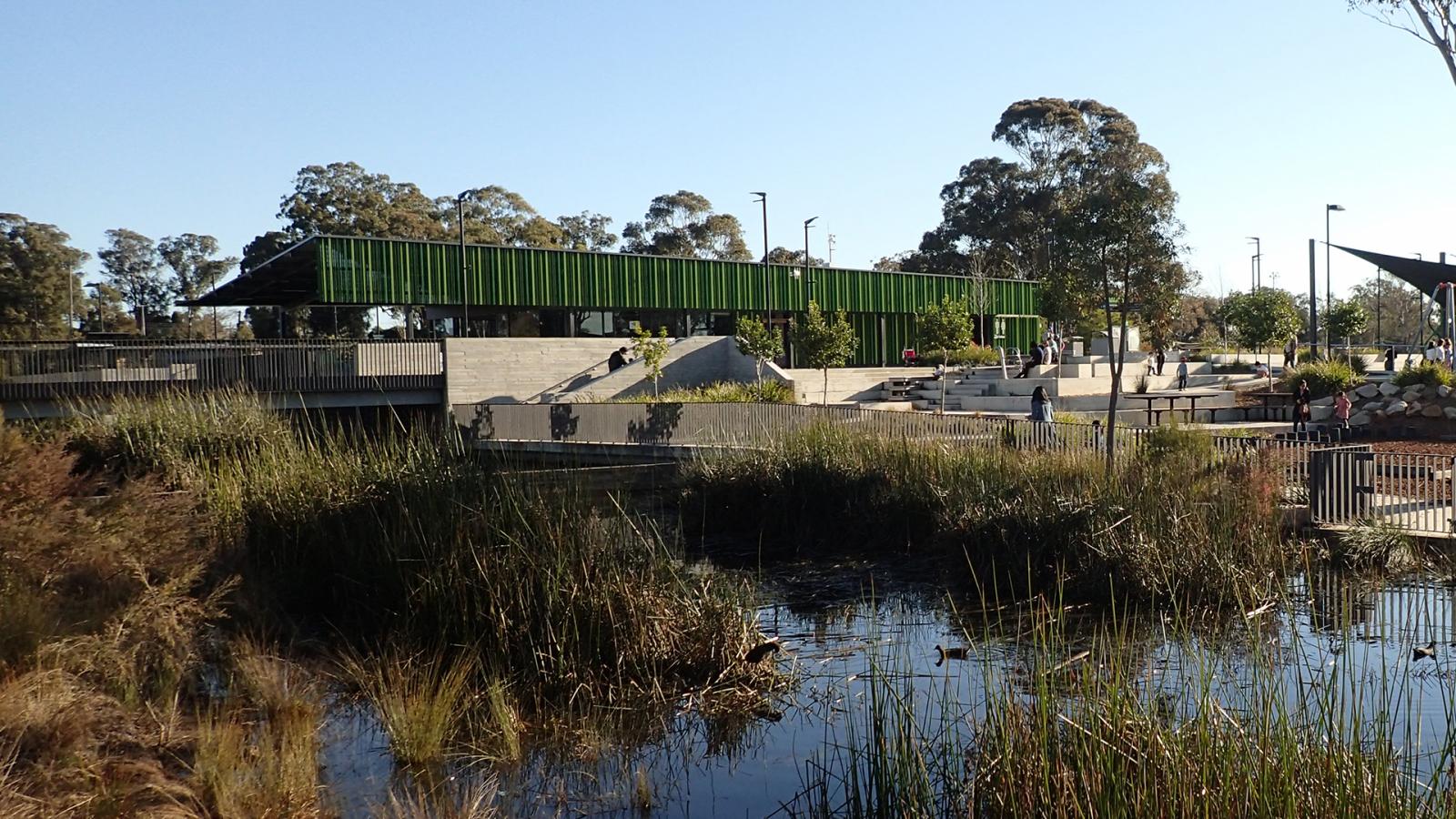 A modern green pavilion with an overhanging roof sits next to a pond with tall grasses in the foreground at the Blacktown showground. The area features concrete steps, paths, and scattered greenery. People can be seen enjoying the outdoor space on a sunny day.