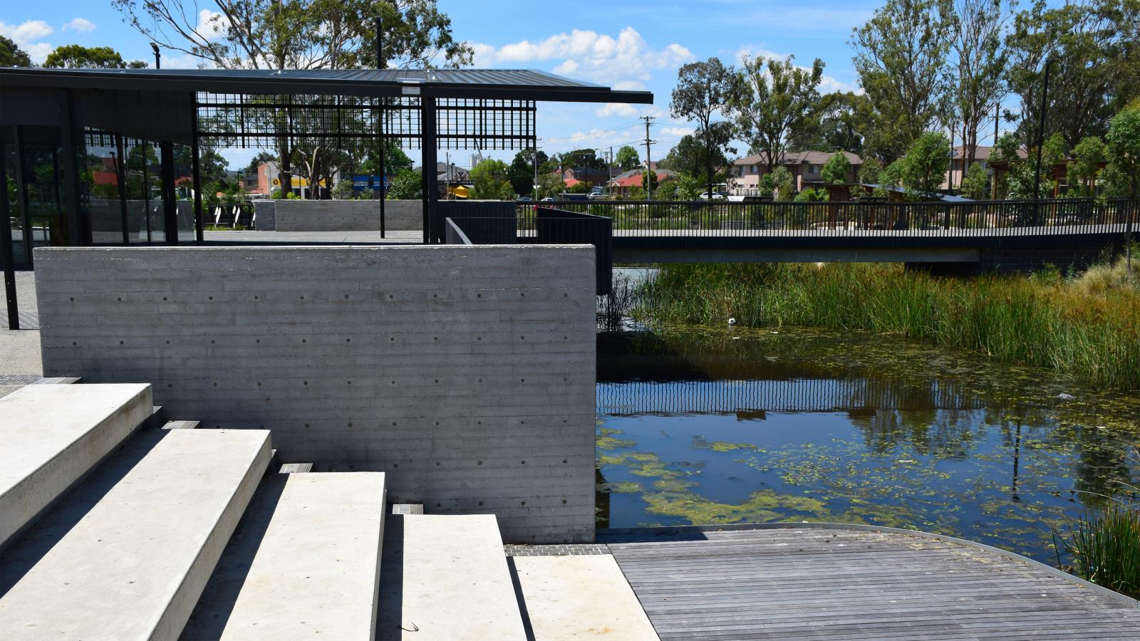 Concrete stairs and a modern pavilion with a lattice-style roof overhang near a water body with reeds and algae, reminiscent of the charming Blacktown Showground. Green trees and residential buildings dot the background, while a bridge spans the water, connecting pathways with metal railings.